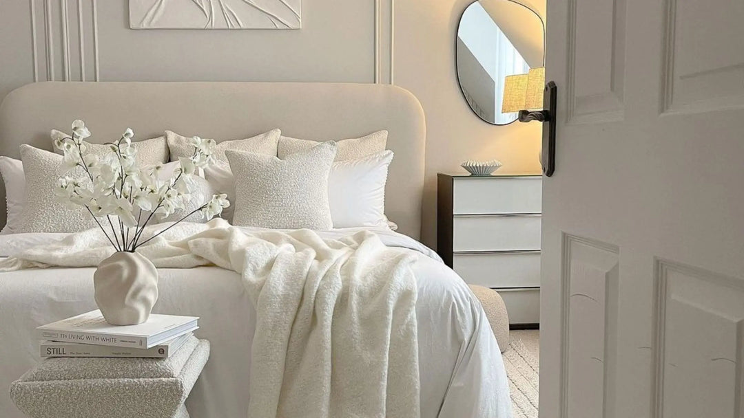 Cream upholstered bed with matching scatter cushions and throw placed on top with small white abstract vase and white flowers on an accent table at the end of the bed