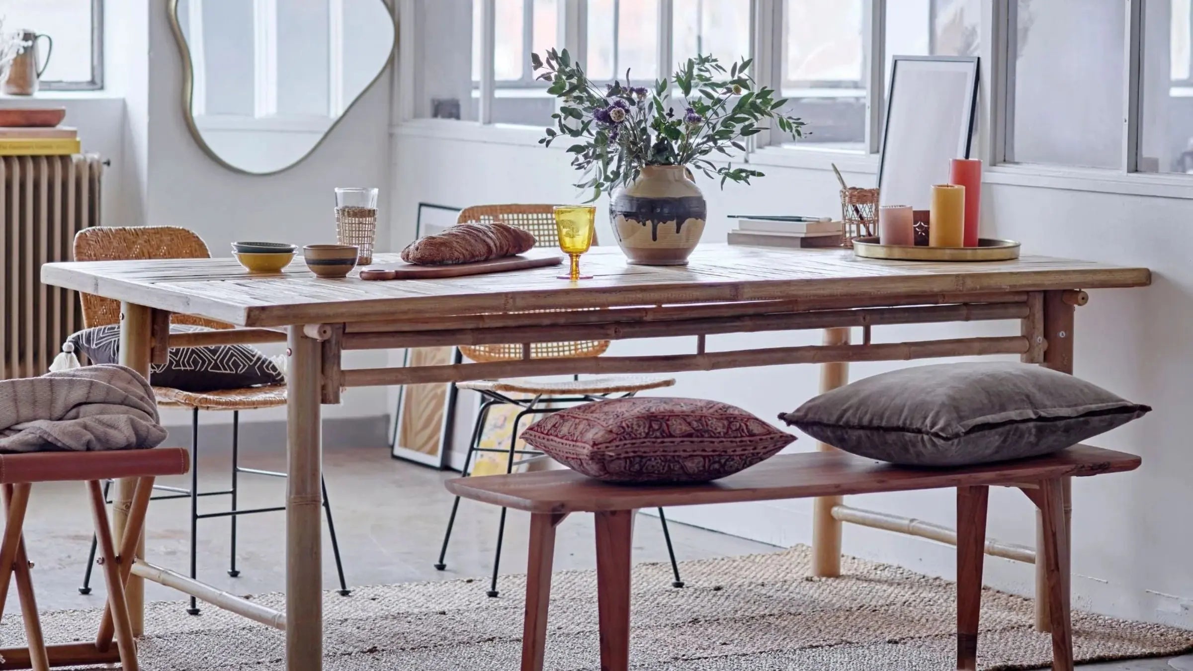 Wooden dining table with wooden bench seating with cushions on, set with colourful glasses, candles and faux foliage