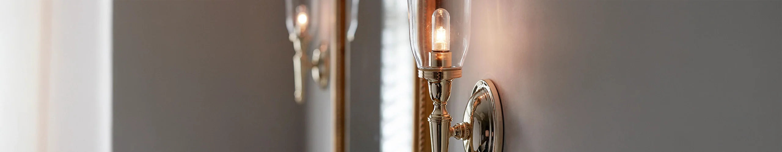 “Elegant brass wall sconce with a cylindrical glass cover, glowing warmly against a smooth gray wall, with its reflection visible nearby.