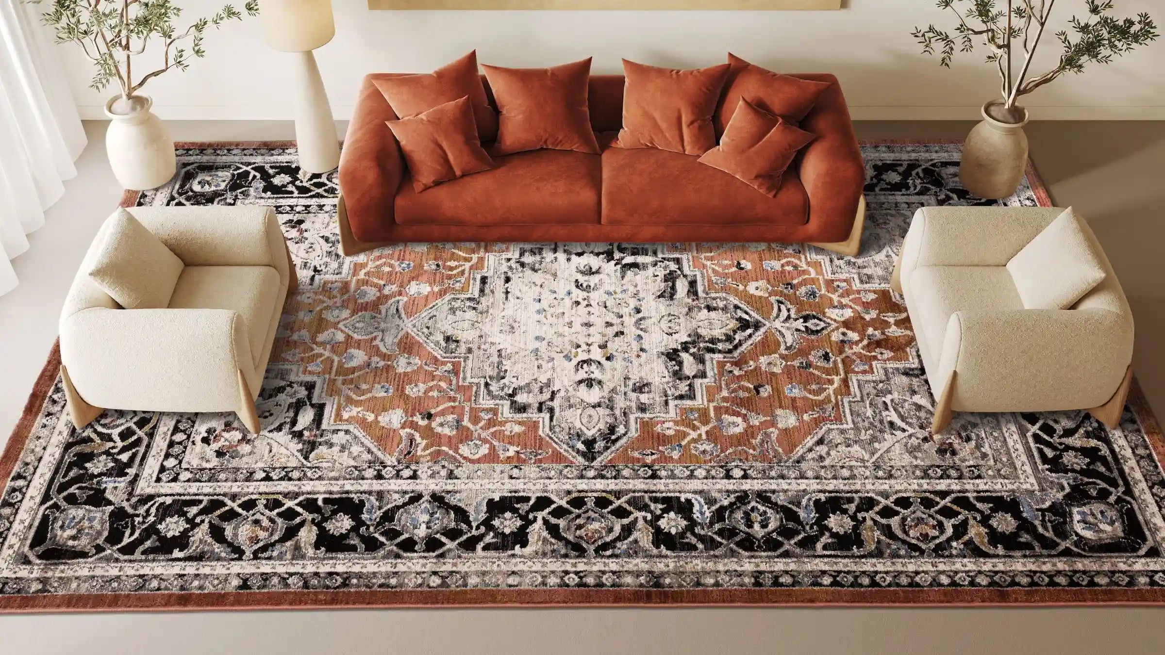 Aerial shot of a living room space with a large patterned rug in the centre, two cream armchairs either end and a rust sofa in the centre with faux plants in stone vases and a cream floor lamp