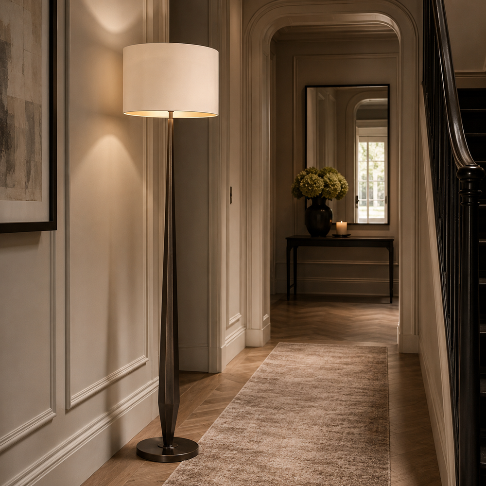 A tall floor lamp with a white drum shade and a dark bronze-toned sculptural base stands in a beautifully lit hallway with wood floors and a neutral rug.