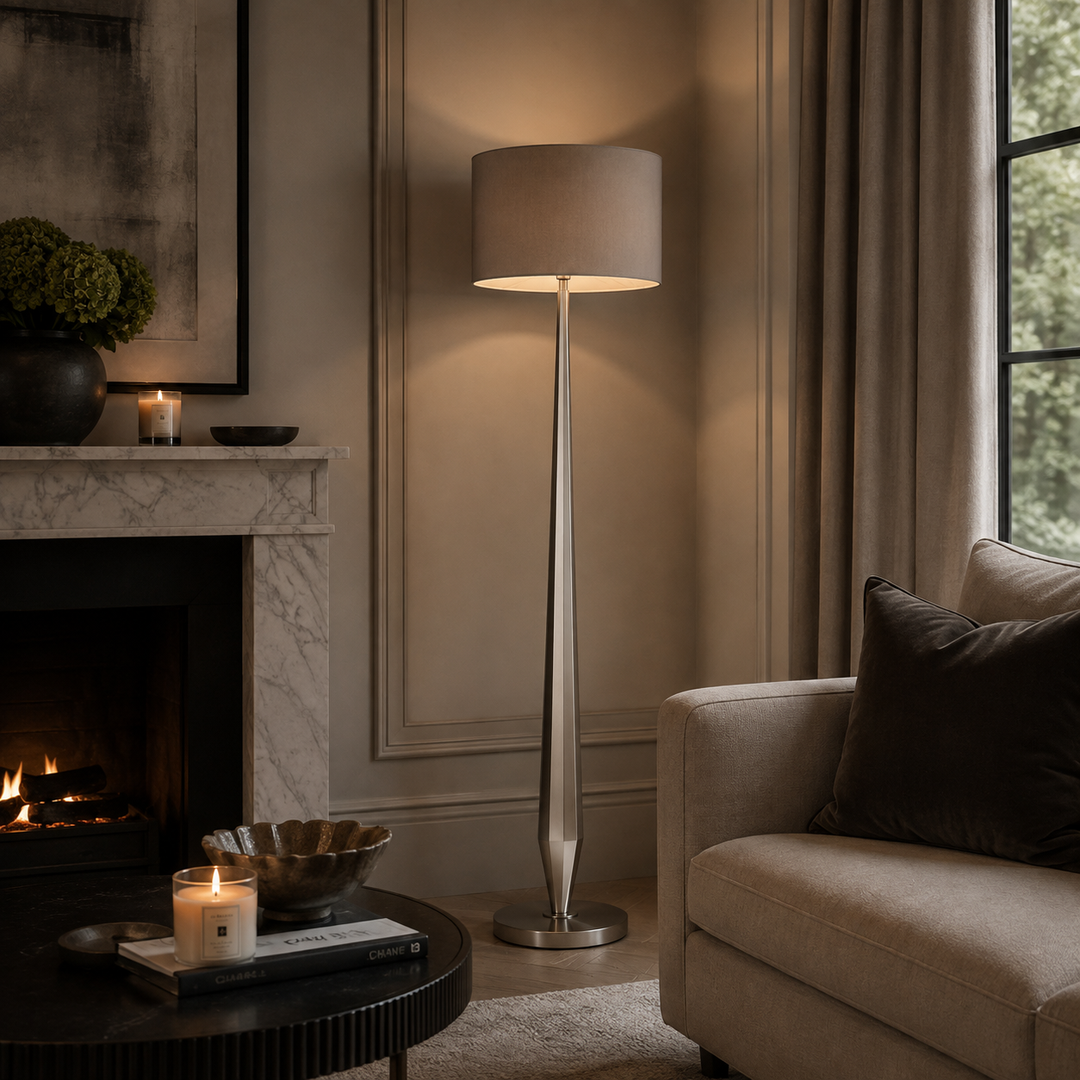 A tall floor lamp with a brushed silver metal stand and a beige fabric drum shade stands in a styled living room next to a fireplace and a beige armchair.