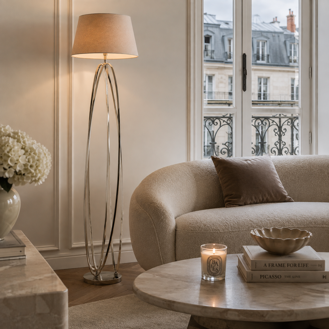 A modern floor lamp with a warm beige fabric drum shade and a polished silver curved metal base stands beside a beige sofa in a softly lit living room. A round marble coffee table, a lit candle, and a vase of white flowers are also visible.