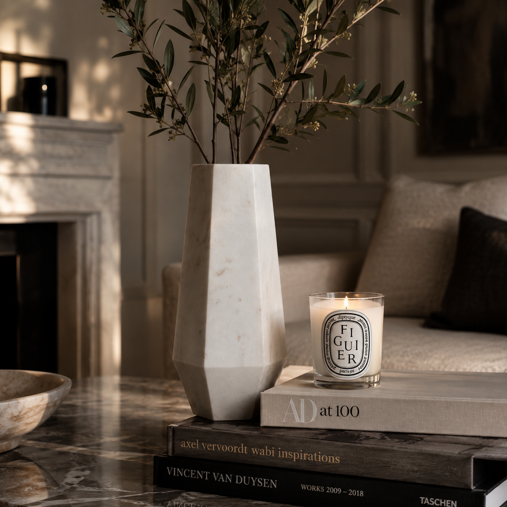 A tall, geometric white marble vase holding olive branches, styled on a marble table next to a lit Diptyque Figuir candle on stacked books in a warm, sunlit living room.