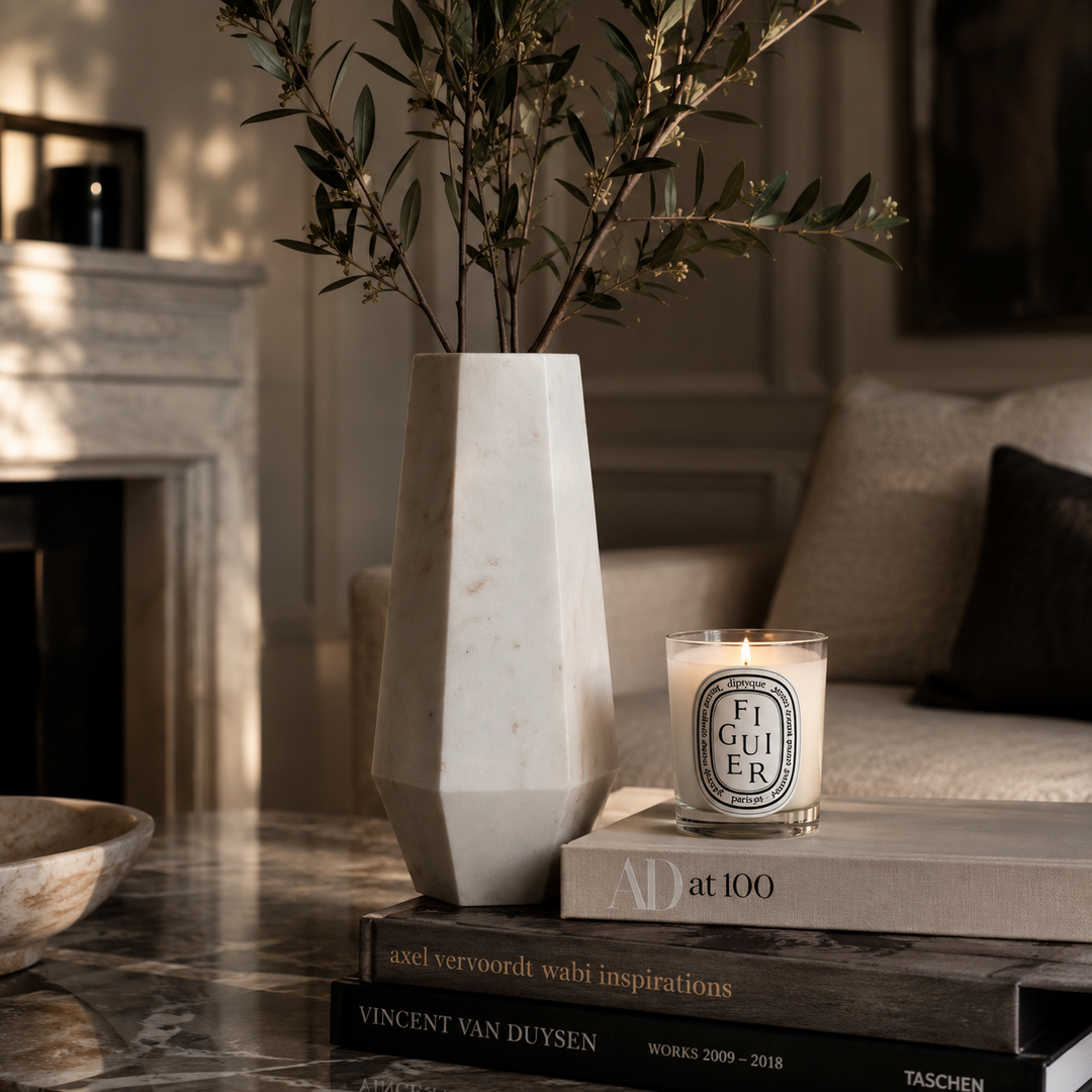 A tall, geometric white marble vase holding olive branches, styled on a marble table next to a lit Diptyque Figuir candle on stacked books in a warm, sunlit living room.