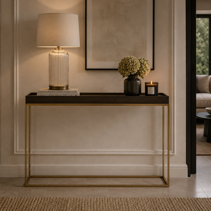 A modern console table with a dark wood top and gold metal frame, styled in a home entryway with a ribbed glass table lamp, a vase of flowers, and a candle.