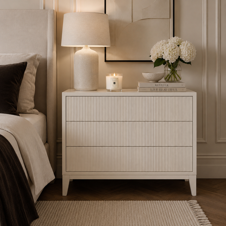 An off-white three-drawer nightstand with a vertical ribbed texture, styled in a bedroom next to a bed with neutral bedding. On top of the nightstand are a lit table lamp, a candle, a small bowl, and a vase of white flowers.