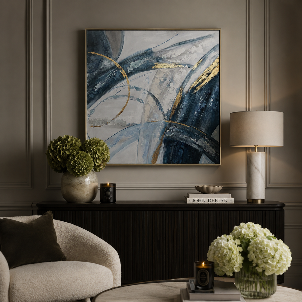 Abstract framed wall art with sweeping navy, blue, gray, white, and gold brushstrokes, displayed above a dark console table in a styled living room with a lamp, flowers, and a chair.