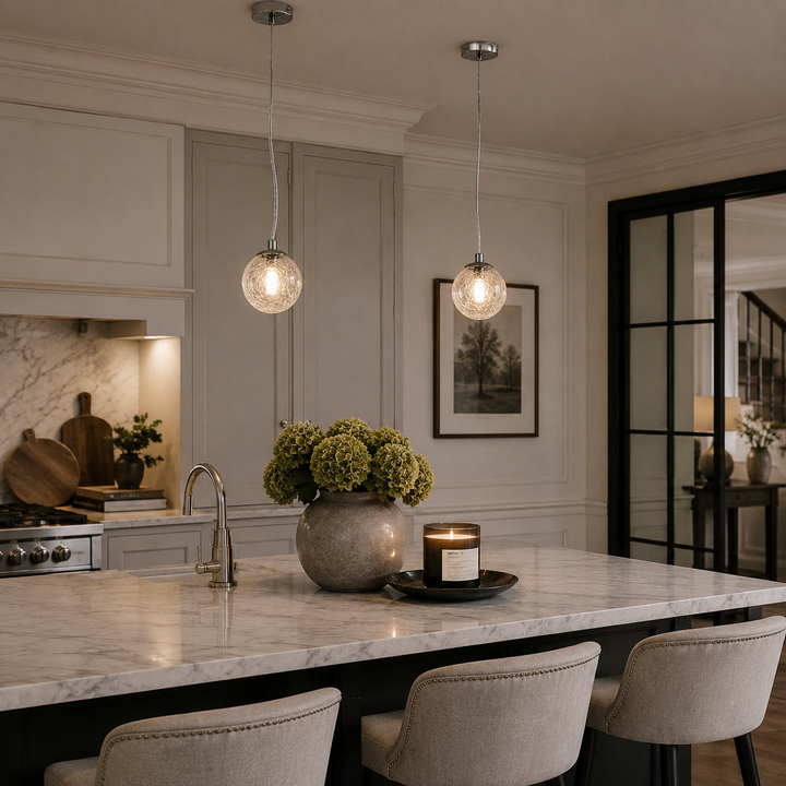 Two spherical glass pendant lights hang over a marble kitchen island in a modern kitchen setting with gray cabinets, a stone vase of greenery, and bar stools.