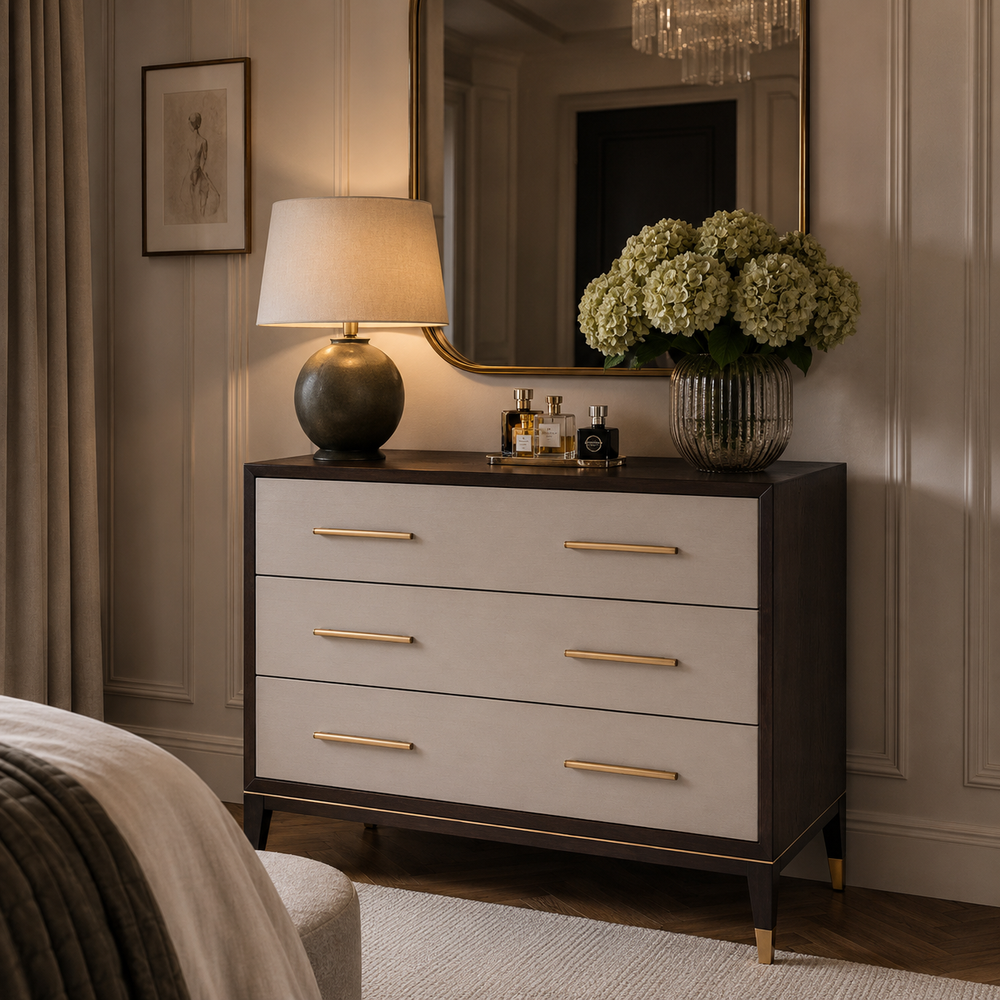 A dark wood and cream dresser in a styled bedroom setting, featuring five drawers with gold handles, a matching lamp, a vase of hydrangeas, and a mirror on top.
