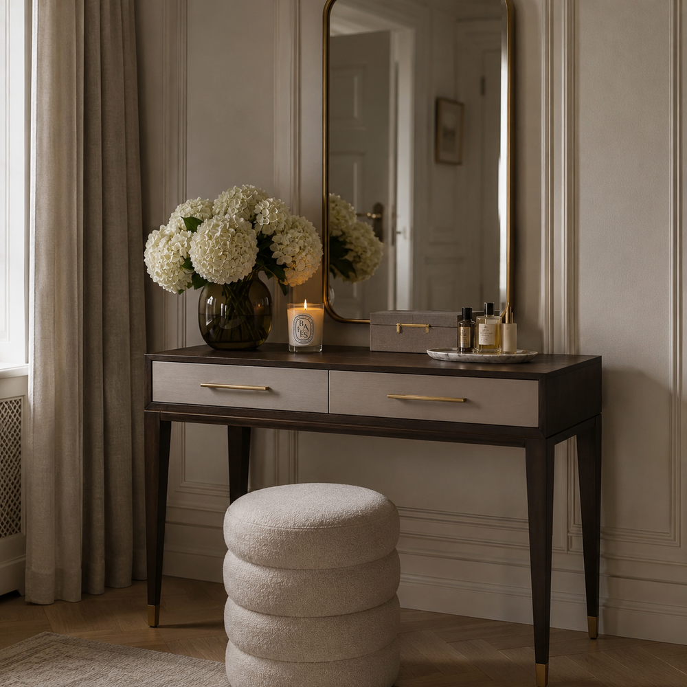 A dark wood vanity table with two drawers and gold-tone handles, styled with a large gold-framed mirror, white flowers, a candle, and toiletries. A round, textured cream ottoman sits in front.