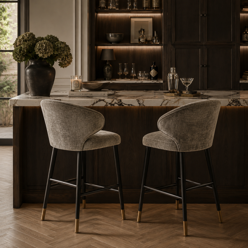 Two upholstered bar stools with curved gray fabric seats and black legs with gold-tone tips, placed at a dark wood kitchen island with a marble countertop in a real interior setting.