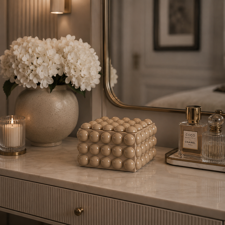 A decorative beige cube-shaped box with a textured spherical bubble pattern sits on a marble vanity table in a styled room setting, next to a vase of white flowers, a candle, and a Chanel perfume bottle.