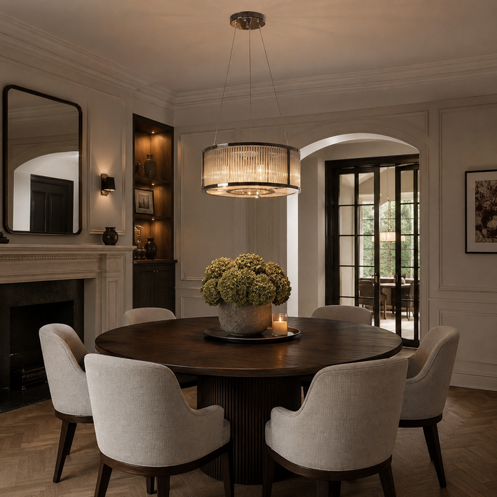 A round dark wood dining table surrounded by six upholstered dining chairs in a warm, elegant dining room. A large cylindrical pendant light hangs above the table, which is decorated with a green hydrangea centerpiece.