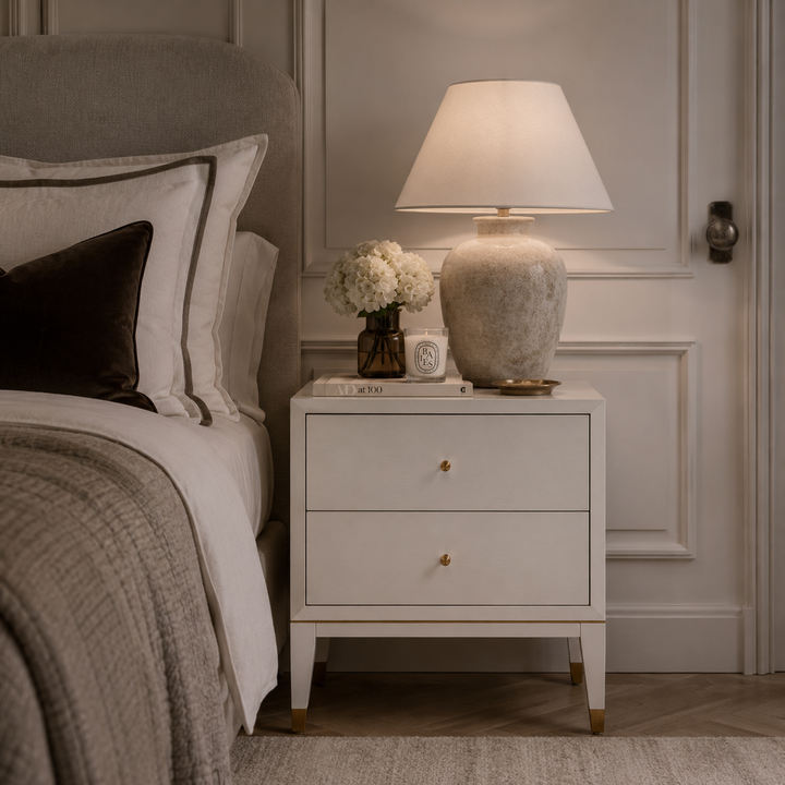 A white two-drawer nightstand in a styled bedroom setting, holding a lit table lamp with a textured ceramic base, a small vase of white flowers, and a clock. The nightstand is positioned next to a bed with neutral-toned bedding and pillows.