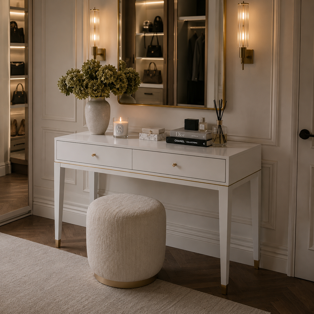 A white vanity desk with two drawers and gold-tone hardware, styled in a luxurious dressing room with a matching round ottoman, a vase of flowers, and a mirror in the background.