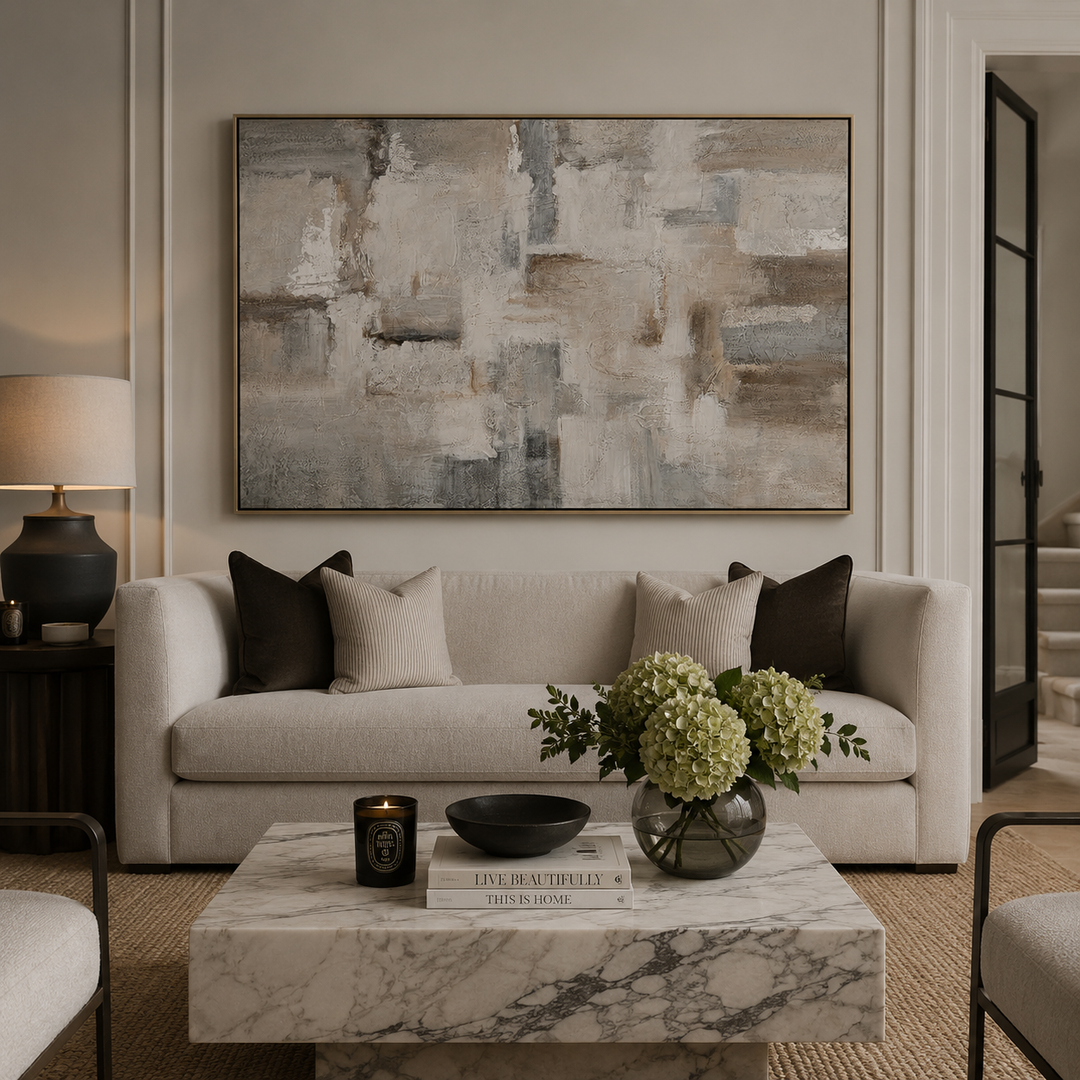 A large framed abstract canvas print with textured brushstrokes in beige, gray, and taupe hangs on a light beige wall above a modern light beige sofa with dark and cream pillows. The scene is styled as a cozy living room with a marble coffee table, a vase of green hydrangeas, and a side table with a lamp.