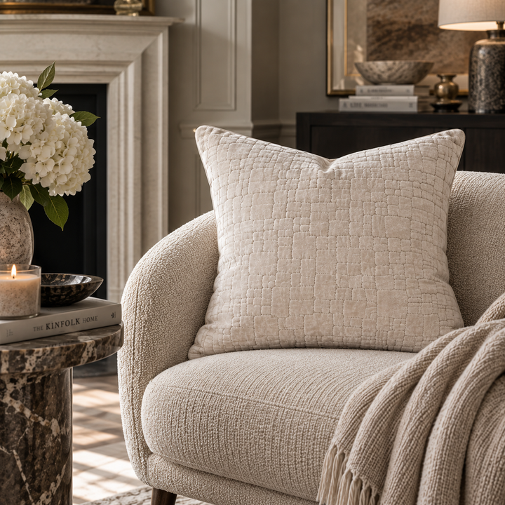 A textured cream throw pillow with a subtle embossed geometric pattern resting on a matching upholstered armchair in a cozy living room setting with a fireplace, candle, and throw blanket.