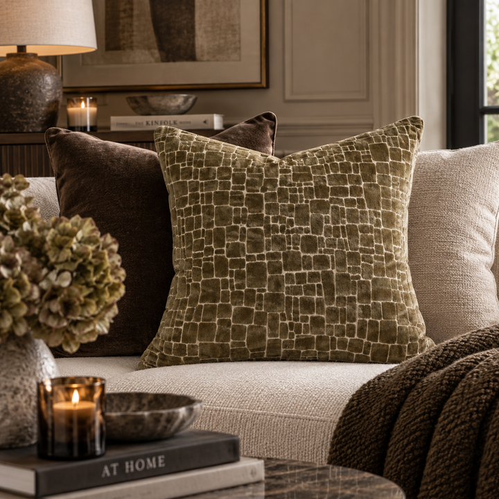 An olive green throw pillow with a raised geometric grid pattern sits on a light beige sofa in a styled living room setting, flanked by a dark brown pillow and a textured brown throw blanket.