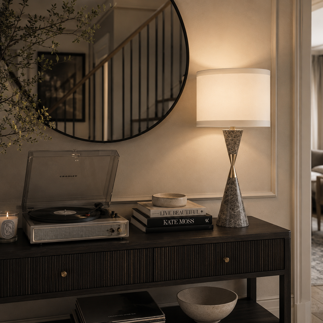 A styled interior scene with a dark wood console table holding a silver turntable, books, a bowl, and a lit table lamp with a white drum shade and textured base. A large round mirror hangs on the wall above, reflecting a staircase, and a doorway is visible on the right.
