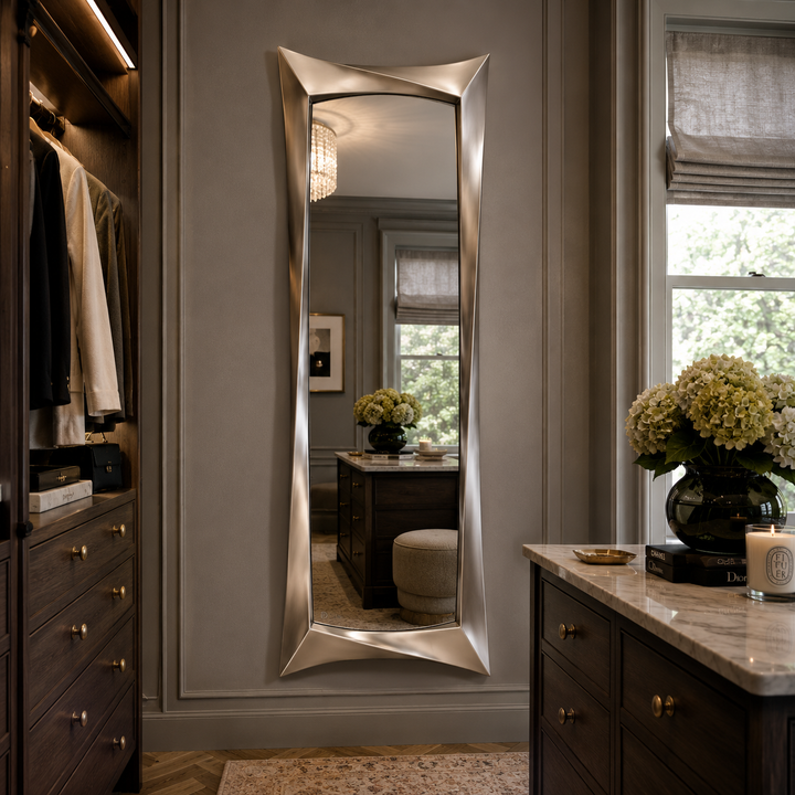 A tall, narrow full-length wall mirror with a sculptural, angular brushed silver frame, mounted on a gray paneled wall in a luxurious dressing room.