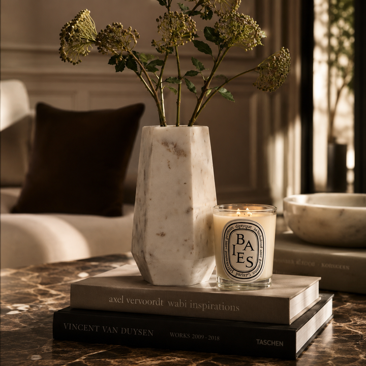 A lit Diptyque Baes candle sits on stacked books beside a white marble vase with dried green stems, styled on a marble tabletop in a softly lit interior setting.