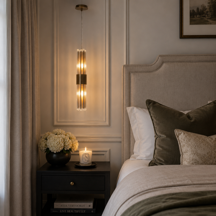 A modern cylindrical wall sconce with a brushed gold finish and warm glowing glass shades, mounted on a beige paneled wall above a bedside table in a styled bedroom.