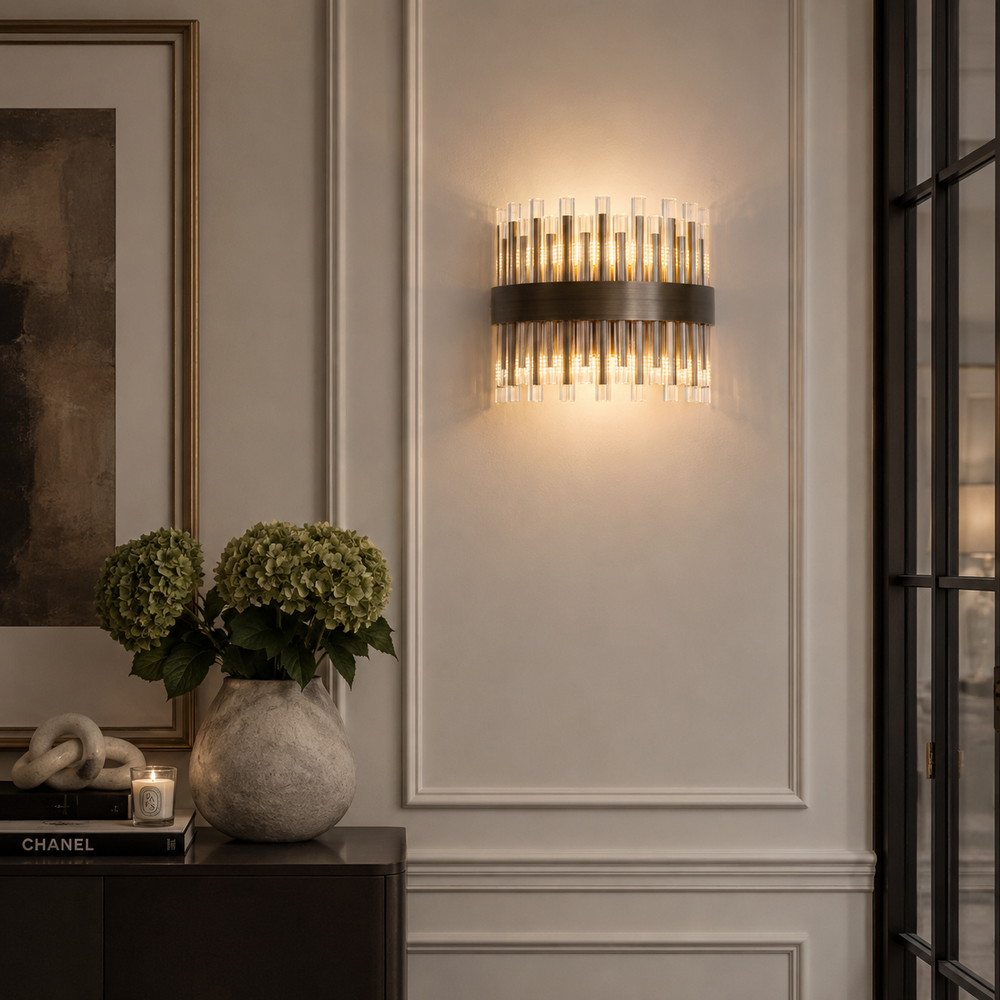 A modern wall sconce light fixture with a dark metal band and multiple clear glass cylindrical tubes, emitting a warm glow on a white paneled wall in an elegant interior setting.