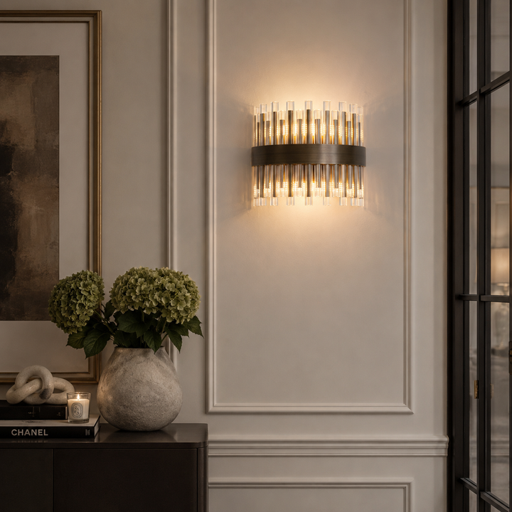 A modern wall sconce light fixture with a dark metal band and multiple clear glass cylindrical tubes, emitting a warm glow on a white paneled wall in an elegant interior setting.