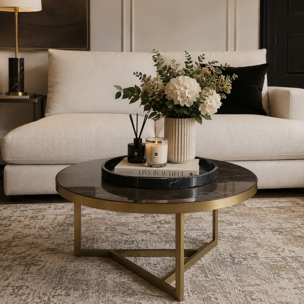 A round coffee table with a black marble top and gold metal legs, styled in a modern living room setting with a cream sofa, floral arrangement, and candles.