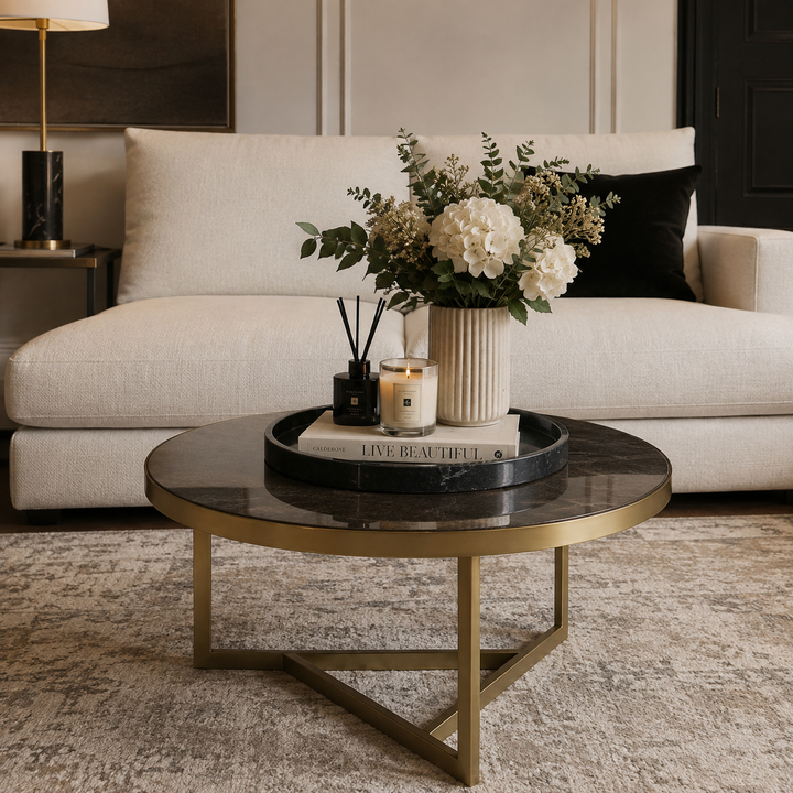 A round coffee table with a black marble top and gold metal legs, styled in a modern living room setting with a cream sofa, floral arrangement, and candles.