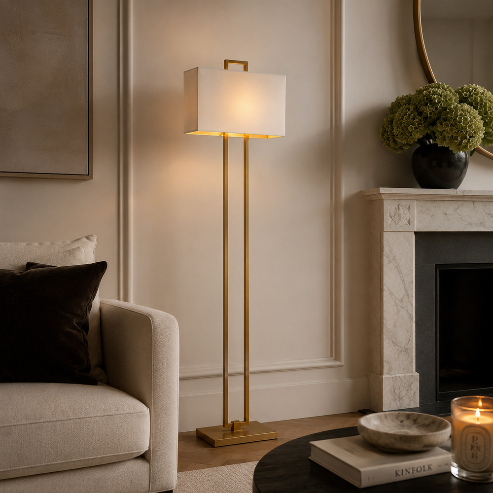 A modern floor lamp with a rectangular white shade and slim gold metal stand, lit in a styled living room beside a sofa and fireplace.