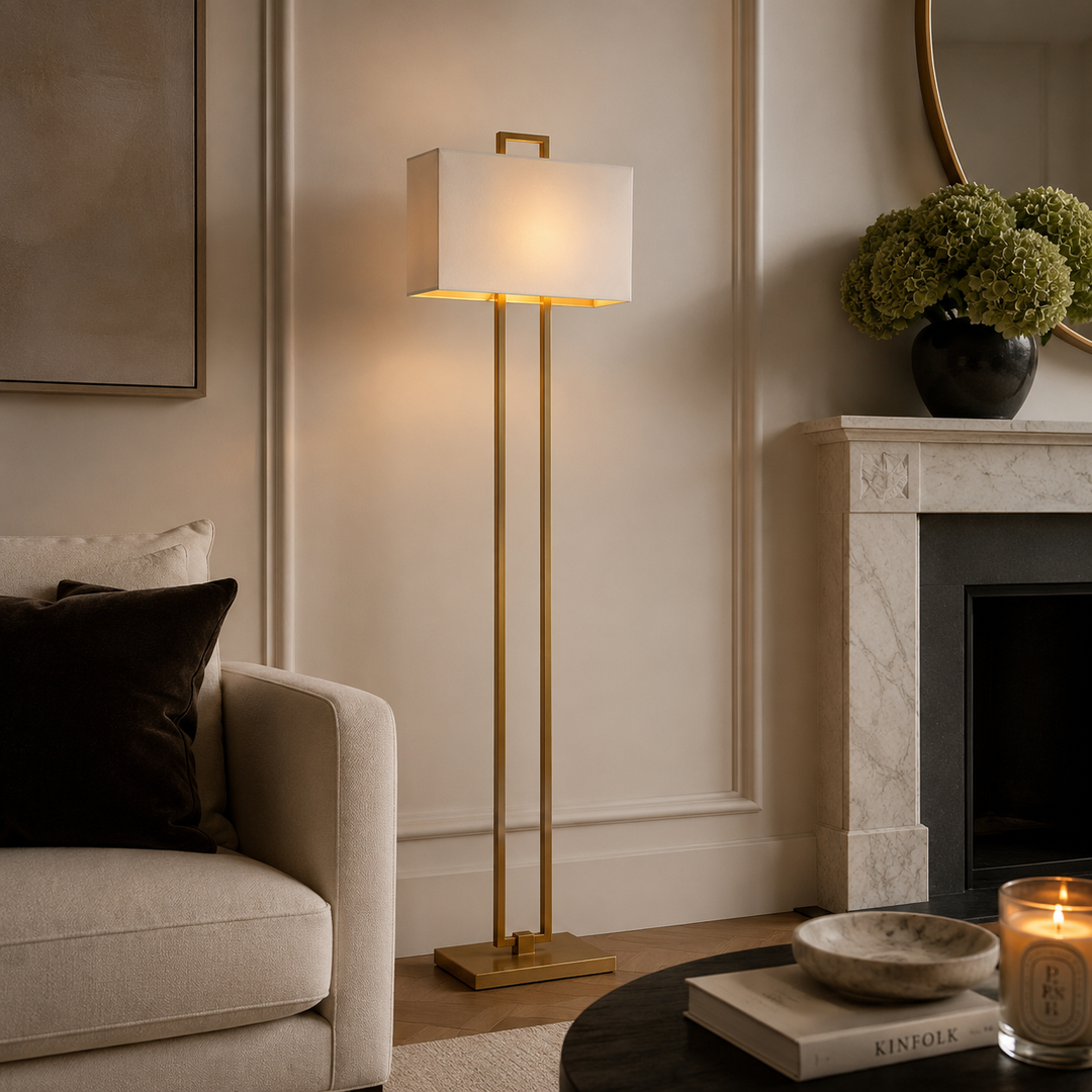 A modern floor lamp with a rectangular white shade and slim gold metal stand, lit in a styled living room beside a sofa and fireplace.