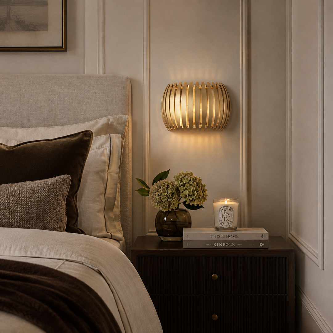 A warm, modern wall sconce with a ribbed cylindrical gold-toned shade glowing on a bedroom wall above a dark wood nightstand.