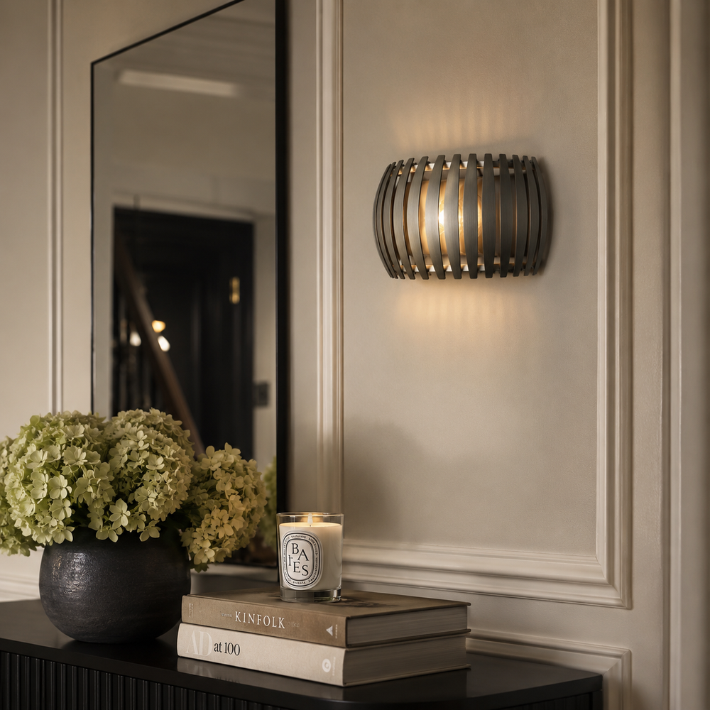 A modern cylindrical wall sconce with a dark metal slatted cage design, emitting a warm glow on a beige paneled wall. Below, a dark console table holds a vase of pale green hydrangeas, a candle, and stacked books, with a large mirror reflecting a dark interior doorway.