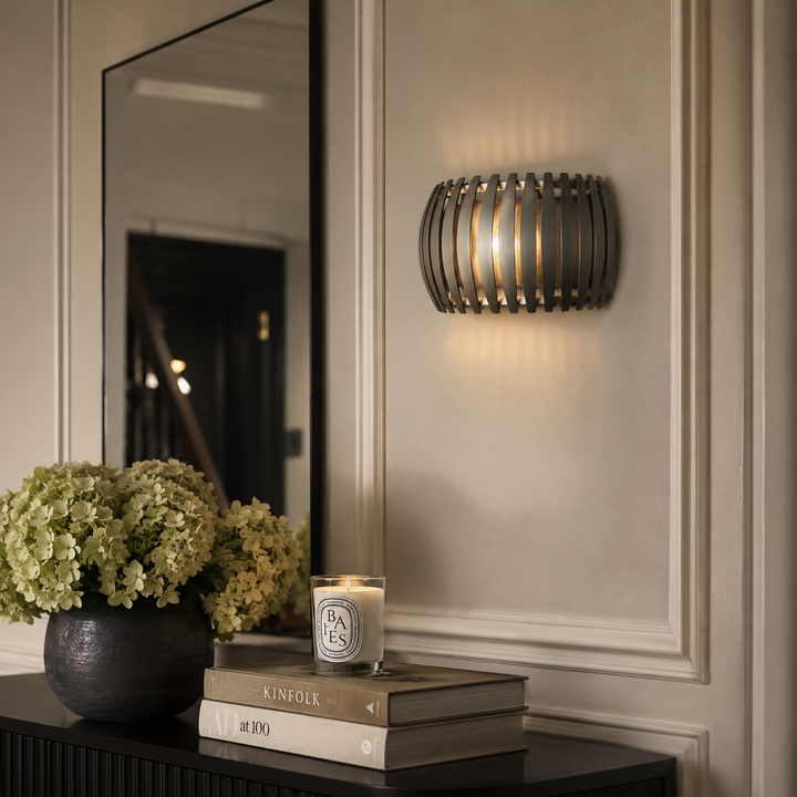 A modern cylindrical wall sconce with a dark metal slatted cage design, emitting a warm glow on a beige paneled wall. Below, a dark console table holds a vase of pale green hydrangeas, a candle, and stacked books, with a large mirror reflecting a dark interior doorway.