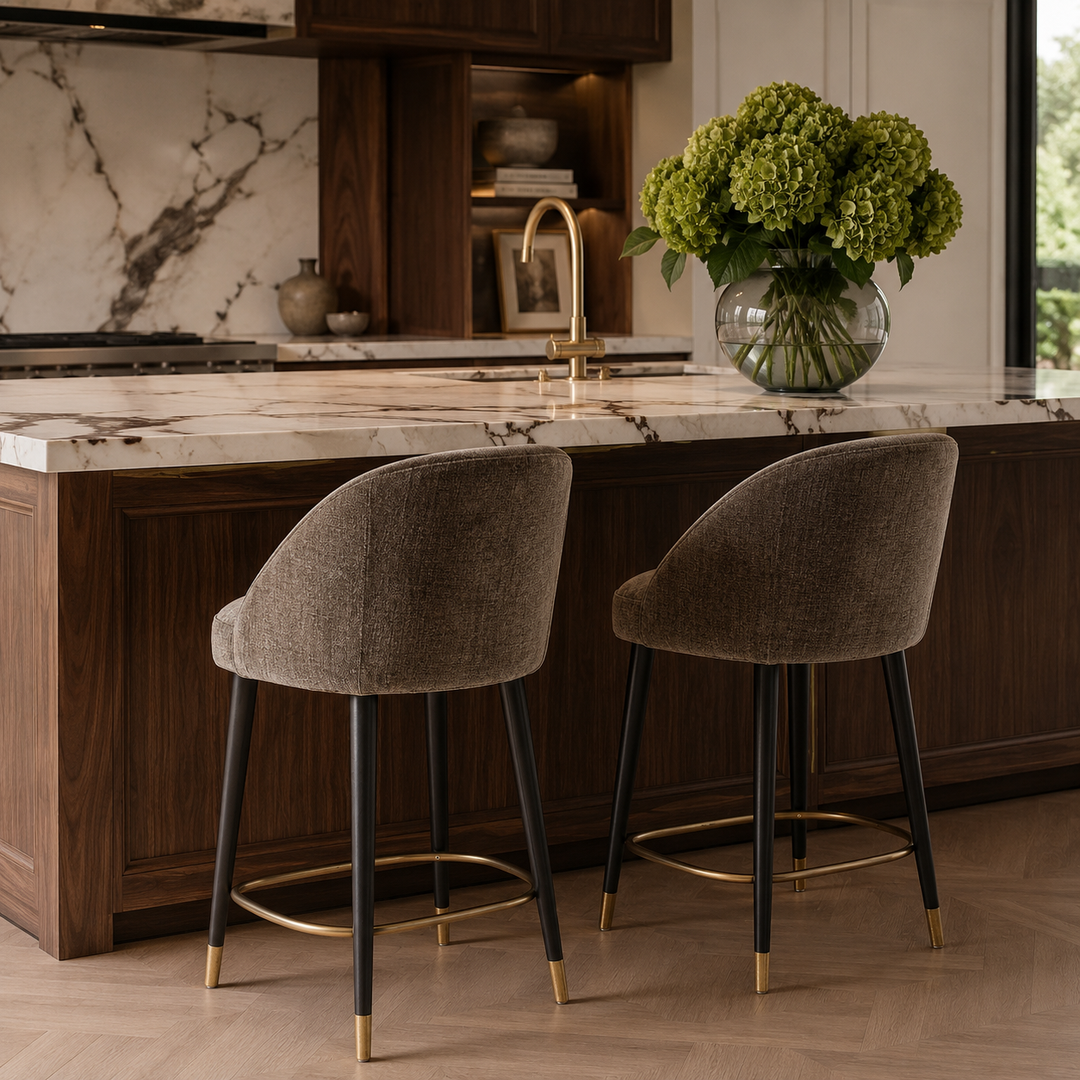 Two upholstered bar stools with taupe fabric seats and black legs with gold-tone feet, placed at a kitchen island with a marble countertop and a vase of green flowers on top.