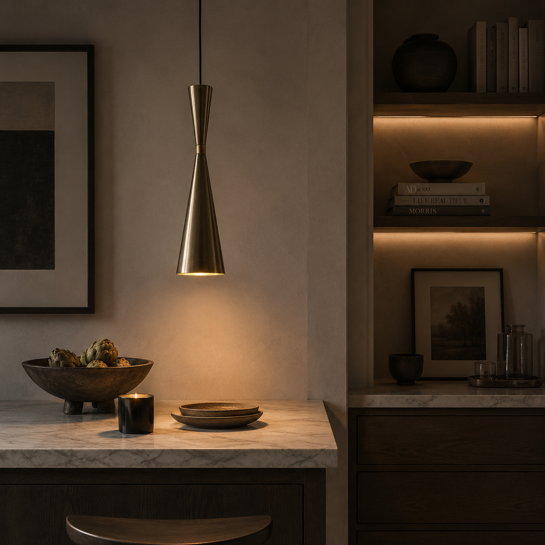 A modern conical pendant light with a brushed brass finish hangs over a marble-topped counter in a dimly lit, stylish interior setting.