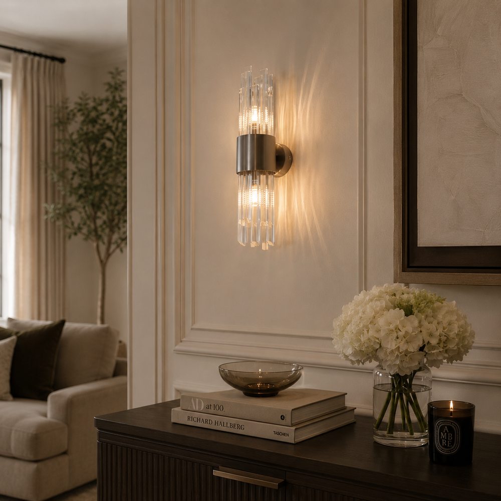 A modern wall sconce with clear glass vertical rods and a brushed brass circular backplate, mounted on a cream paneled wall above a dark wood console table with books, a glass bowl, and a vase of white flowers.
