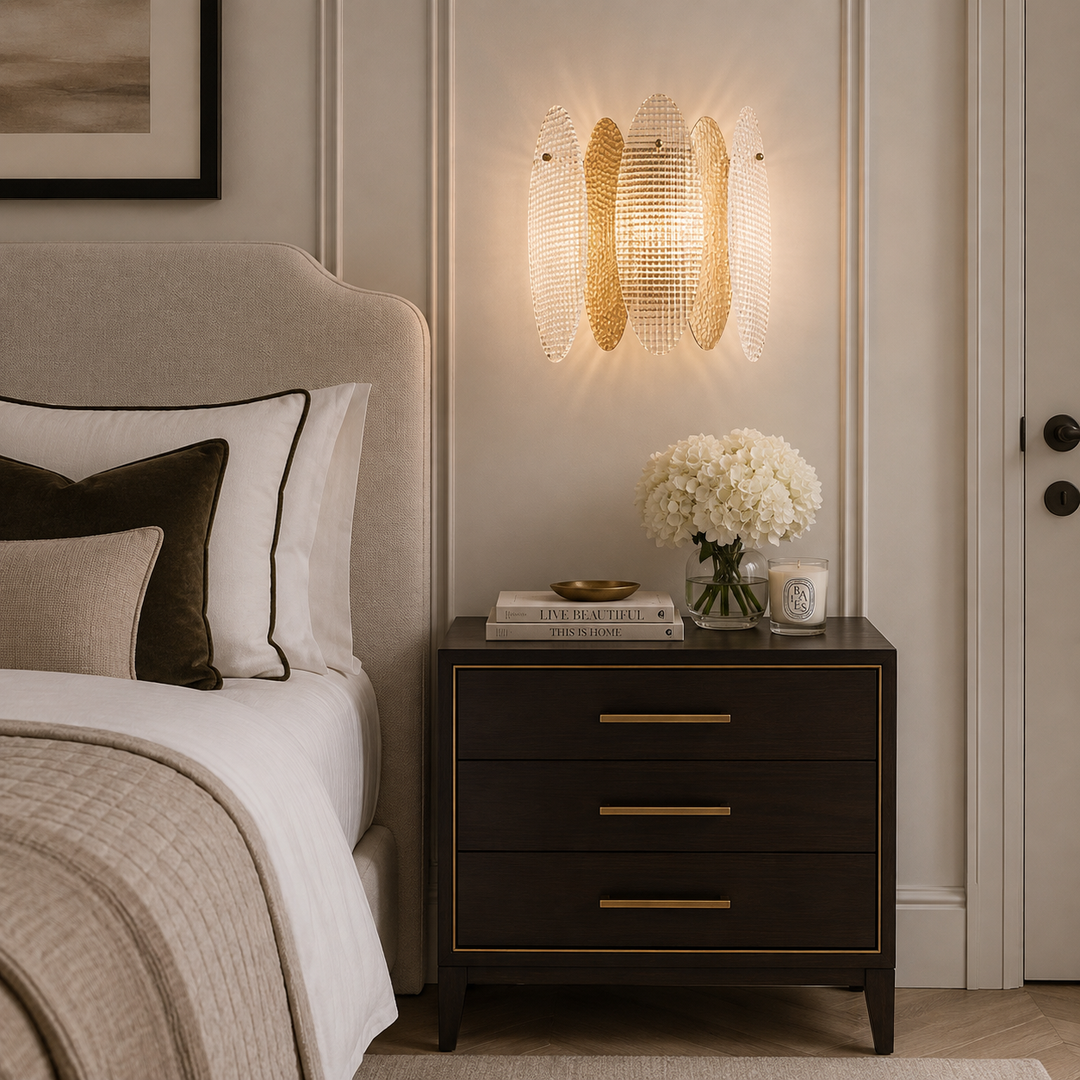 A modern wall-mounted light fixture with three curved gold-toned glass panels glowing warm light, installed above a dark wood nightstand in a styled bedroom setting.
