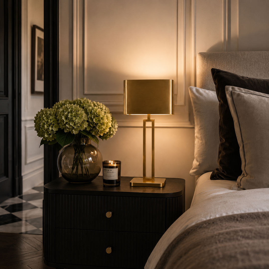 A modern table lamp with a rectangular gold-toned shade and matching base sits on a dark bedside table next to a bed. The scene is warmly lit, with a vase of pale green hydrangeas and a candle on the table, set in an elegant bedroom interior.