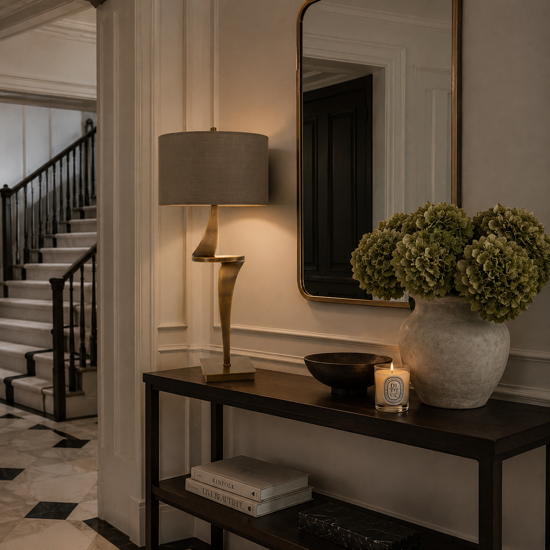 A styled interior entryway scene with a dark wood console table holding a lit table lamp, a ceramic vase of green hydrangeas, a bowl, a candle, and books. A large mirror hangs on the wall, and a staircase is visible in the background.