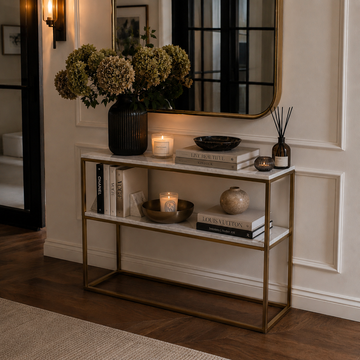 A modern two-tier console table with a gold metal frame and white marble shelves, styled in a warm, elegant entryway with books, candles, a vase of hydrangeas, and a mirror.