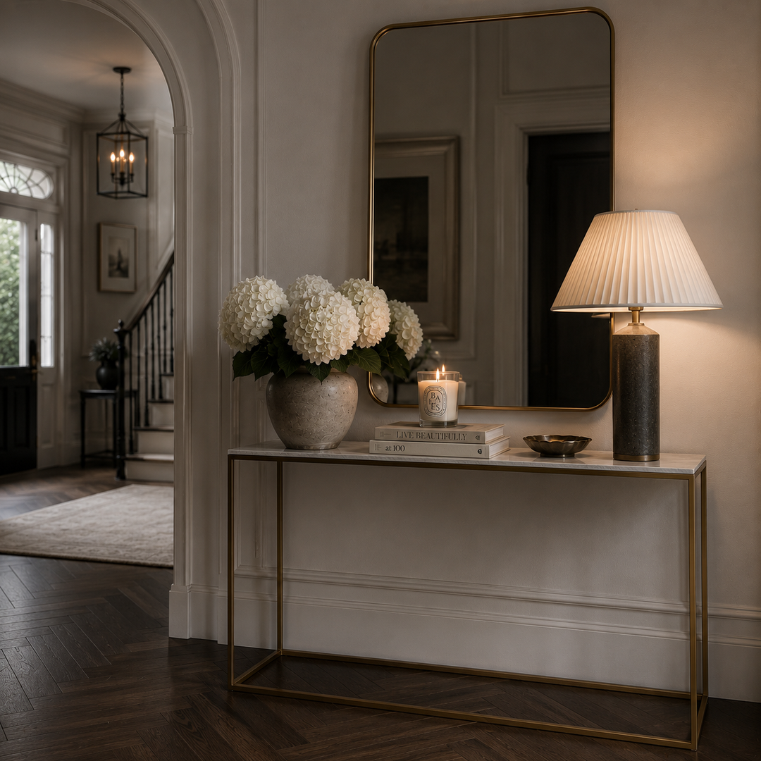 A modern console table with a gold metal frame and white marble top, styled in a classic entryway. On top of the table are white hydrangeas in a textured vase, a lit candle, a book, and a small bowl.