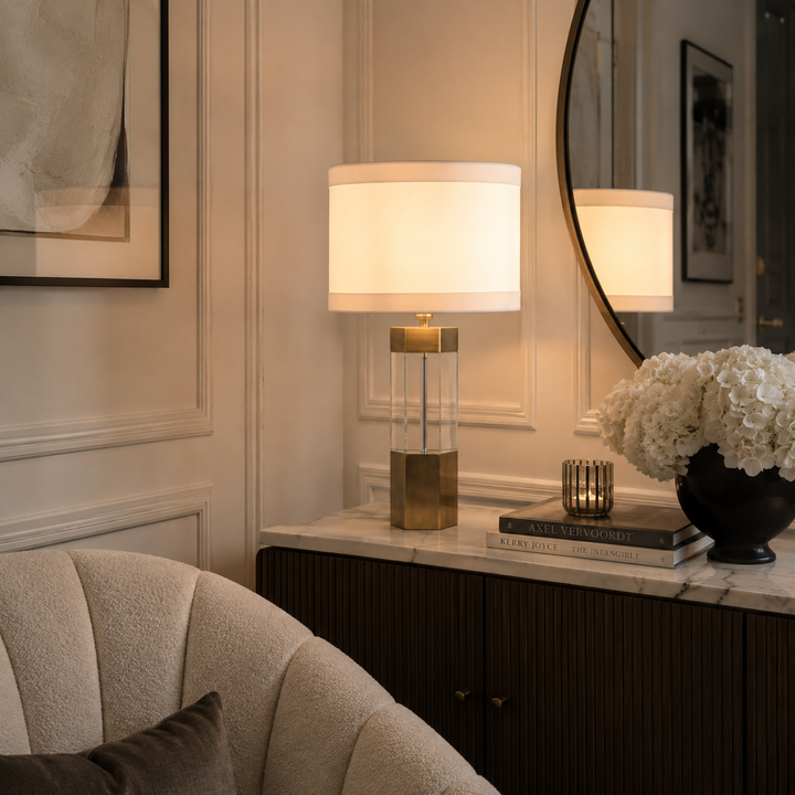 A table lamp with a warm white drum shade and a gold-and-clear geometric base sits on a marble console table in a styled interior. A round mirror, framed wall art, books, a candle, and a vase of white flowers are visible around it.