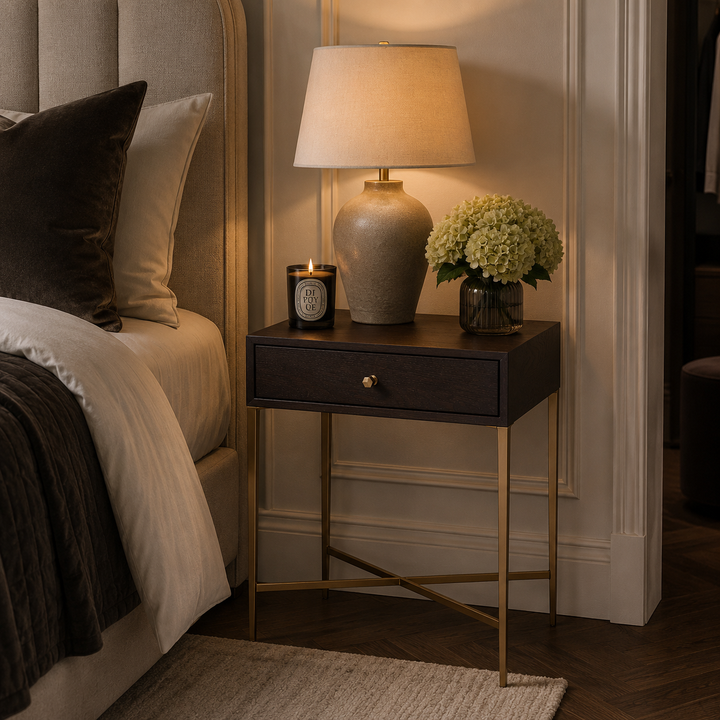 A dark wood nightstand with a single drawer and brass legs in a bedroom setting, styled with a lit table lamp, a candle, and a vase of green hydrangeas next to a bed.