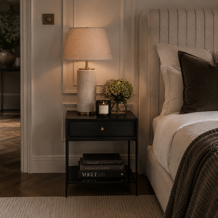A bedside table in a styled bedroom setting, featuring a lit table lamp with a beige fabric shade and textured base, a candle jar, a small floral arrangement, and stacked books on a black metal frame table next to a bed with neutral-toned bedding.