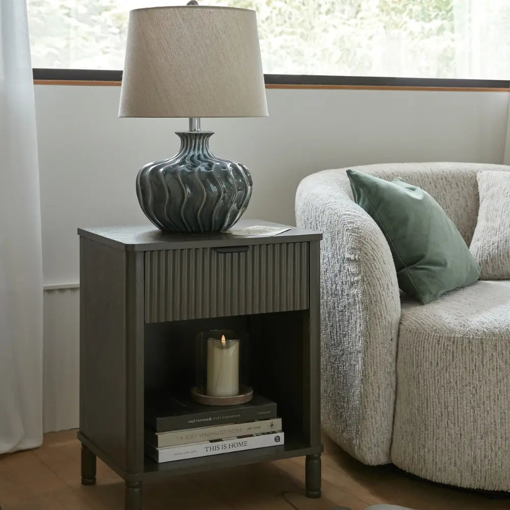 Gray side table with lamp and books next to a beige sofa with a green pillow.