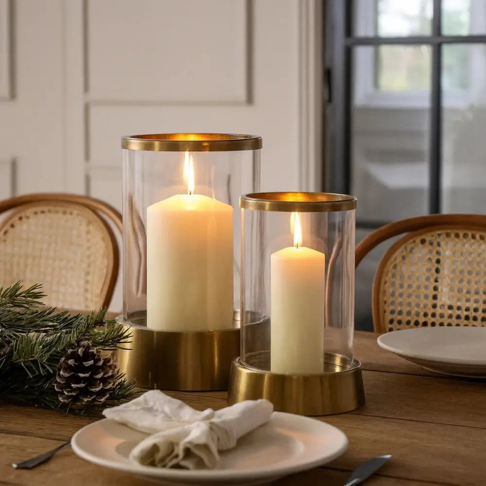 Two lit candles in gold holders on a table with plates and greenery.