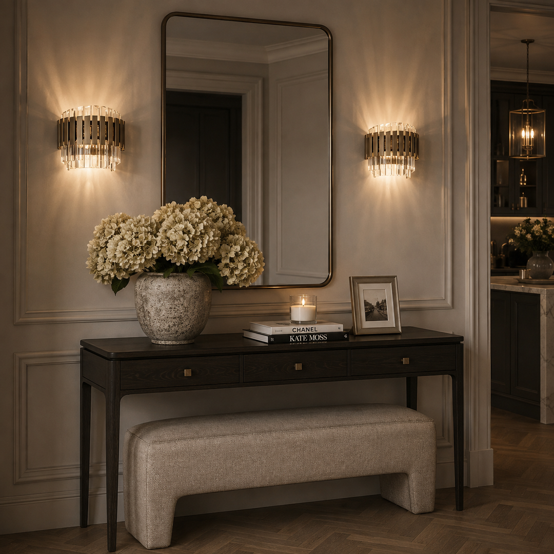 A styled interior scene with a dark wood console table, upholstered bench, large wall mirror, and two illuminated cylindrical wall sconces. A vase of cream flowers, books, and framed art sit on the table against a paneled wall.